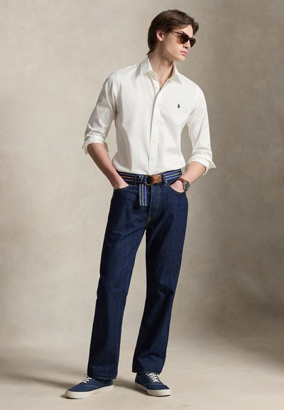 Νεαρός άνδρας που φοράει λευκό πουκάμισο με κουμπιά, μπλε ζώνη με ρίγες, σκούρο τζιν, μπλε αθλητικά παπούτσια και γυαλιά ηλίου, στέκεται με τα χέρια στις τσέπες.