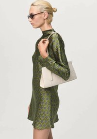 Vestido verde con estampado, de cuello alto y mangas largas; sostiene un bolso crema con diseño angular. El atuendo parece elegante y moderno.
