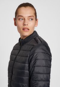 Giacca nera, morbida e gonfia con colletto alto e dettagli di cucitura orizzontale. Chiusura con zip sul davanti. Tessuto di texture liscia e lucida.