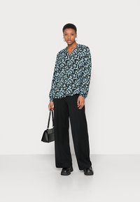 Blusa floral preta com flores azuis, mangas longas, decote em V. Combinada com calças largas pretas e uma pequena mala preta.