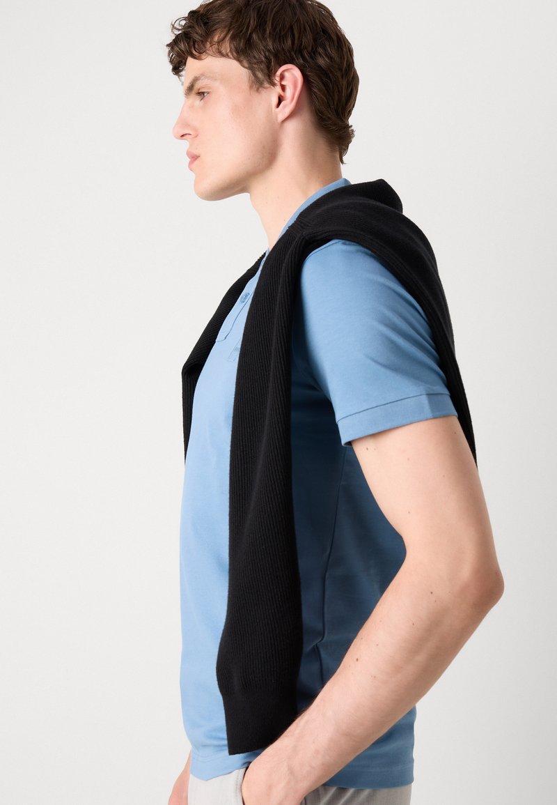 Camisa polo de color azul claro con un suéter negro de canalé caído sobre los hombros, que presenta un collar con botones y mangas cortas.