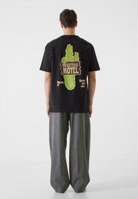 Melnas īsām piedurknēm T-krekli ar zaļu kaktusu drukājumu un tekstu "WESTERN MOTEL" mugurpusē; viss apvienots ar pelēkām plašām biksēm.