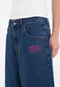 Dark blue denim jeans with a loose fit, featuring a pink embroidered logo on the left pocket and a classic five-pocket design.