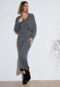 Vestido de punto gris con canales y un cárdigan corto que presenta botones dorados. Botines negros de tobillo completan el look sobre un fondo sencillo.