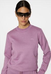 Felpa di cotone viola con collo rotondo e polsini a coste, con un sottile accento del logo. Il modello indossa occhiali da sole scuri.