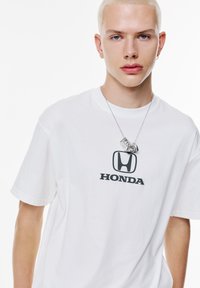 Hvid bomulds t-shirt med et sort "HONDA" logo trykt på fronten. Modellen bærer en sølv halskæde med abstrakt vedhæng.