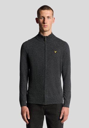 Sweater zippé gris foncé avec un col montant, des poignets côtelés et un petit emblème jaune sur la poitrine, fabriqué en matériau tricoté doux.