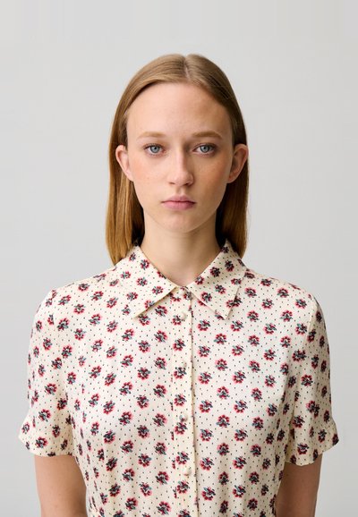 Giovane donna con capelli biondi lisci che indossa una camicia a maniche corte color crema con motivo floreale rosso, che guarda direttamente avanti con espressione neutra.