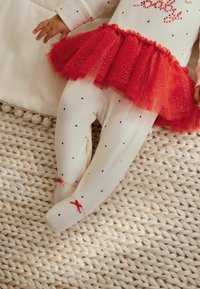 Hvid bomuldsbody med rød tylskørt, prydet med små sorte hjerter. Har røde sløjfer ved anklerne. Sat mod en struktureret vævet baggrund.