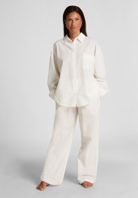 Witte button-up shirt met een zak en een gestructureerd patroon, gecombineerd met bijpassende loszittende broek van een zachte stof, dit alles zonder schoenen.