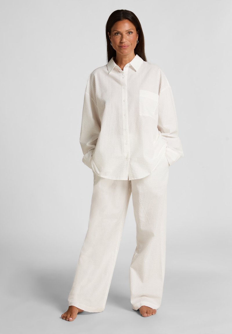 Witte button-up shirt met een zak en een gestructureerd patroon, gecombineerd met bijpassende loszittende broek van een zachte stof, dit alles zonder schoenen.