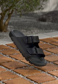 Sandale noire texturée avec deux sangles à boucle réglables flottant au-dessus d'un sol en carreaux de bois et en cailloux, avec un arbre flou en arrière-plan.