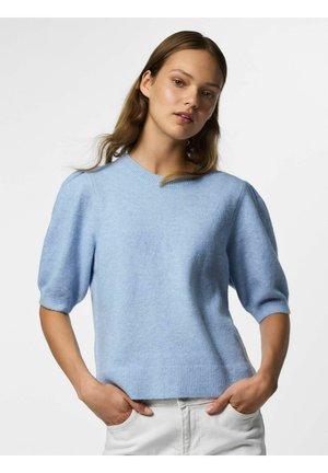 Femme aux longs cheveux bruns portant un pull bleu clair à manches courtes et un pantalon blanc, debout les mains dans les poches devant un fond uni.