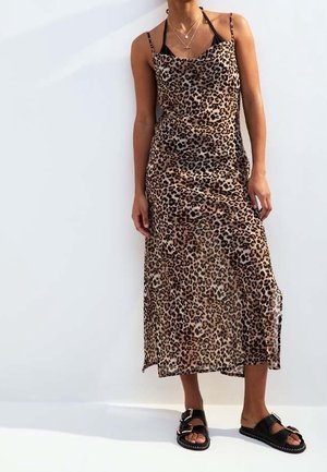 Nainen pukeutuneena hihattomaan leopardikuvioiseen midihameeseen, mustat sandaalit jalassa ja kerrokselliset kultaiset kaulakorut, seisomassa valkoisen taustan edessä.