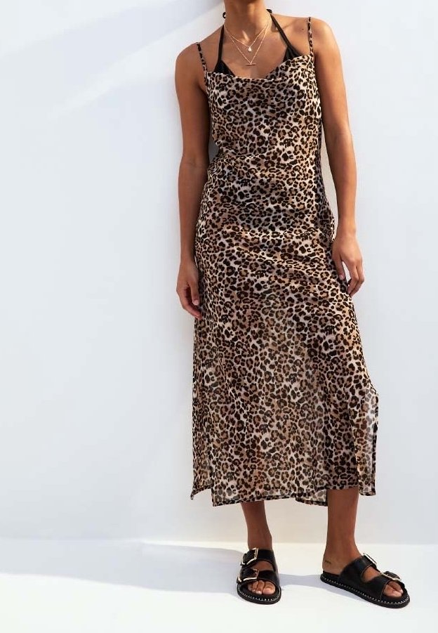 Femme portant une robe midi sans manches à imprimé léopard, avec des sandales noires et des colliers en or superposés, debout devant un fond blanc.