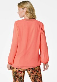 Korallenfarbene Bluse mit langen Ärmeln, die einen gerafften Ausschnitt, eine lockere Passform und eine durchsichtige Textur aufweist, kombiniert mit gemusterten, mehrfarbigen Hosen.