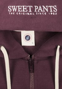 Sweat à capuche violet avec un cordon blanc et une fermeture éclair, orné de texte brodé "SWEET PANTS" et d'une étiquette affichant les détails de la marque.