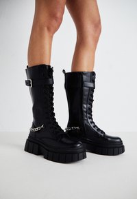 Schwarze knielange Kampfstiefel mit Schnürung an der Vorderseite, chunky Plateausohle, Reißverschluss und einem silbernen Kettenakzent an der Seite.