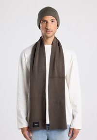 Bufanda de punto acanalada marrón y gorro a juego, llevados con una camiseta de manga larga blanca. La bufanda cuenta con una pequeña etiqueta de logo en el dobladillo.