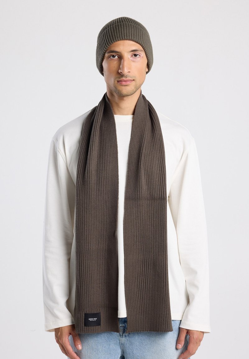 Bufanda de punto acanalada marrón y gorro a juego, llevados con una camiseta de manga larga blanca. La bufanda cuenta con una pequeña etiqueta de logo en el dobladillo.