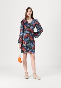 Robe florale noire avec des motifs vibrants rouges, bleus et verts, dotée de longues manches bouffantes, d'un décolleté en V et d'une silhouette ajustée. Sac à main orange.