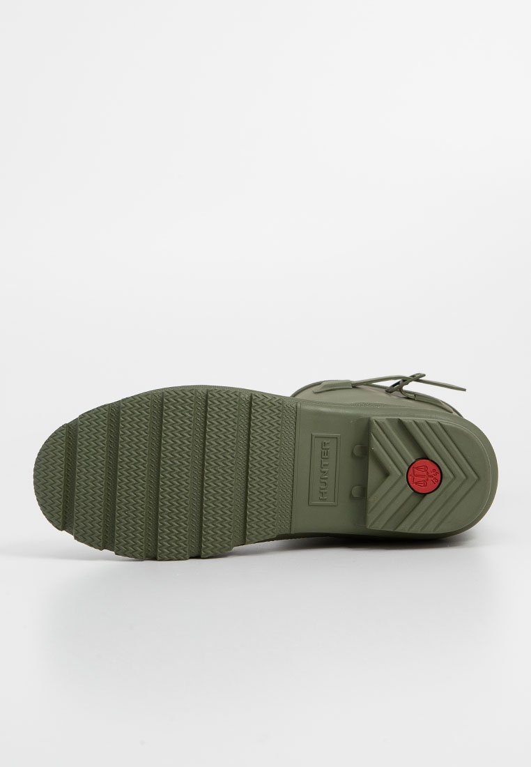 Bota de goma verde oliva con suela texturizada y estriada. Presenta una punta redondeada, diseño de cordones y un acento de logo rojo cerca del talón.