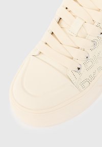 Suure plaaniga kreemika värvusega tennis обувь, paksude nööridega, tekstuuritud tallaga ja perforatsioonidega külgpaneelidega, millel on õrn muster.