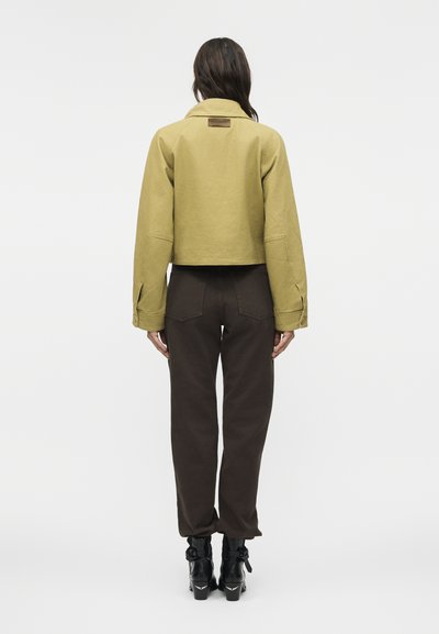 Γυναίκα που στέκεται με το πρόσωπο προς τα πίσω, φοράει μια κοντή ελιά μπουφάν, σκούρο καφέ παντελόνια και μαύρα μποτάκια αστραγάλου με αγκράφες σε απλό λευκό φόντο.