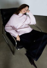Lys rosa striktrøje med ribbet manchetter, parret med vidde denim bukser i mørk farve. Model siddende i en elegant sort læderstol.