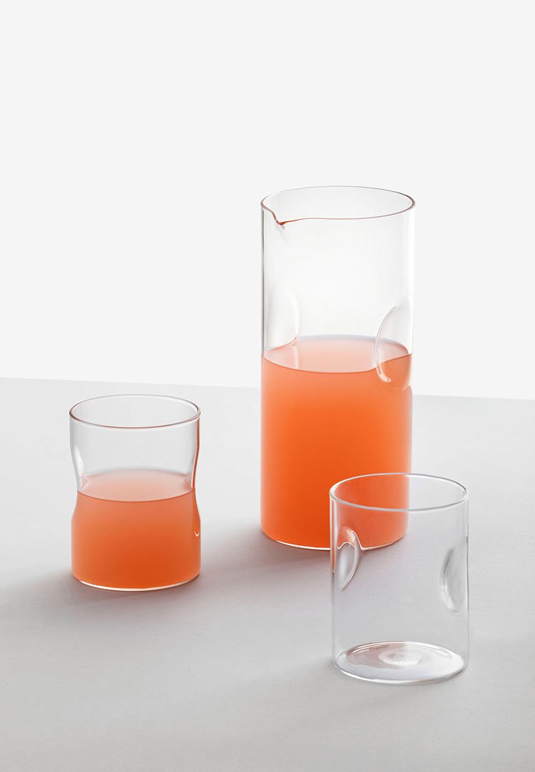 Trzy przezroczyste szklane naczynia: wysoki dzbanek i dwa krótsze kubki, wszystkie zawierające różową ciecz. Elementy konstrukcyjne obejmują zaokrąglone boki i gładkie krawędzie.