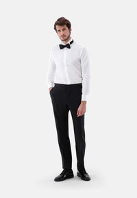 Weißes Hemd mit schwarzer Fliege, schwarze Hose und glänzenden schwarzen Schuhen. Das Outfit ist im taillierten Schnitt und hat eine glatte Textur.