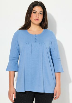 Lichtblauwe, langslavige shirt met een ronde halslijn, plooien en knoopdetails op de borst. Zachte stof met een ontspannen pasvorm.