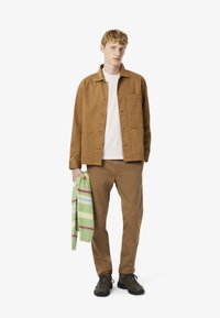 Veste en coton marron avec deux poches avant, portée sur une chemise rose clair, assortie à un pantalon marron et des chaussures noires, tenant un pull rayé multicolore vert.