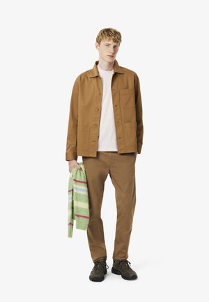 Giacca di cotone marrone con due tasche frontali, indossata sopra una camicia rosa chiaro, abbinata a pantaloni marroni coordinati e scarpe nere, mentre tiene un maglione verde a righe multicolori.