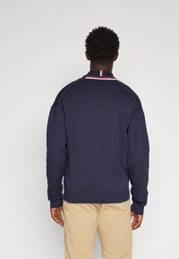 Marineblauer Strickpullover mit strukturiertem Rippenmuster, ausgestattet mit einem rot-weißen gestreiften Kragen und kontrastierenden Schulterakzenten. Lockere Passform.