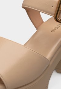 Μπεζ δερμάτινο σανδάλι ανοιχτού δακτύλου με λουράκι στον αστράγαλο και χοντρό τακούνι, με χρυσή αγκράφα και ορατό όνομα μάρκας στην εσωτερική σόλα.