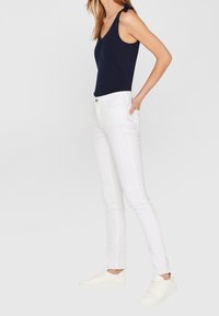 Marinefarbener ärmelloser Bodysuit kombiniert mit weißen, eng anliegenden Jeans und weißen Sneakers. Der Bodysuit hat einen Rundhalsausschnitt, und die Jeans verfügen über einen Reißverschluss und einen Knopf.