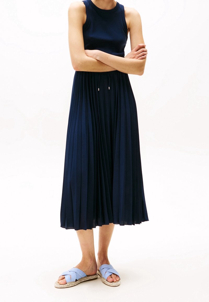 Femme portant une robe midi plissée sans manches bleu marine et des sandales slip-on bleu clair, debout, les bras croisés.