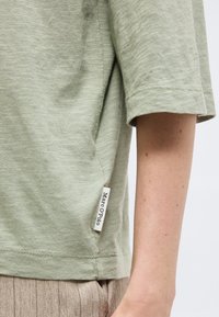 Ανοιχτό πράσινο κοντομάνικο πουκάμισο με ετικέτα «Marc O'Polo» στην αριστερή πλευρά, φορεμένο πάνω από καφέ υφασμάτινο παντελόνι, με μερική προβολή χεριού και μέσης.