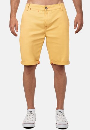 Mann in gelben hochgekrempelten Shorts und weißen Turnschuhen steht vor einfachem Hintergrund.