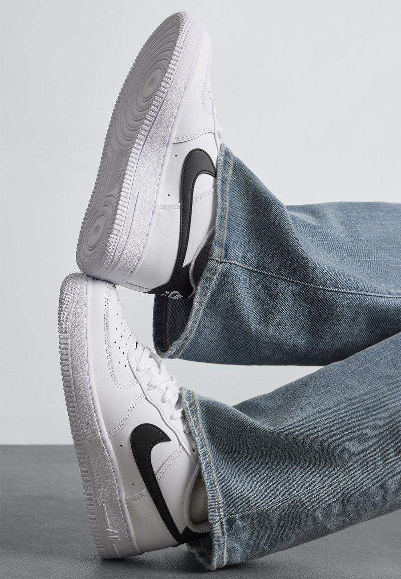 Baskets blanches avec le logo noir Nike, portées avec un jean bleu sur un fond gris clair uni.