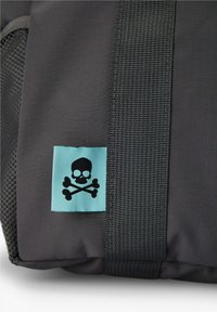 Tela negra y gris con una correa negra gruesa y una etiqueta azul que muestra un símbolo de calavera y huesos cruzados en negro.