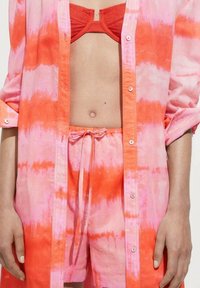 Ein Batik-T-Shirt in Pink und Orange, mit einem Button-Up-Design und hochgekrempelten Ärmeln, kombiniert mit passenden Batik-Shorts und einem roten Bikini-Oberteil.