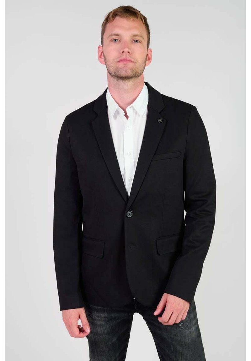 Zwarte blazer met twee voorzakken en enkele knoopsluiting, gedragen over een wit shirt, in combinatie met donkere jeans. Gladde stofstructuur.