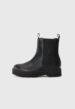 Botas Chelsea de cuero negro con punta redonda, paneles laterales elásticos, lengüetas para tirar y una suela de goma gruesa con un dibujo pronunciado.