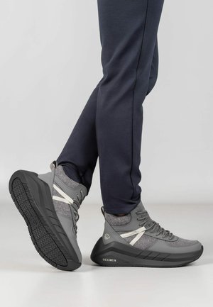 Para szarych i czarnych wysokich sneakersów z białymi akcentami noszona z ciemnymi spodniami, jedna stopa oparta na pięcie, pokazująca wzór bieżnika podeszwy.