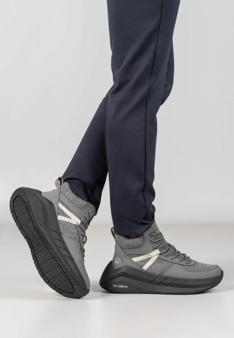 Paar graue und schwarze High-Top-Sneaker mit weißen Akzenten, getragen zu dunkler Hose, ein Fuß ruht auf der Ferse und zeigt das Profil der Sohle.