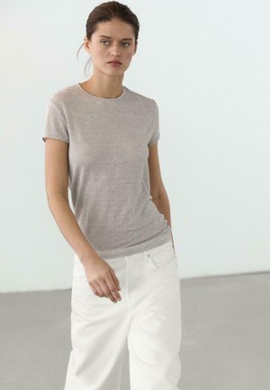 Giovane donna che indossa una maglietta a maniche corte grigio chiaro e pantaloni bianchi larghi, in piedi davanti a uno sfondo bianco uniforme.