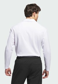 Vit långärmad pikétröja med krage, kombinerad med svarta byxor med elastisk midja. Slätt tyg och figursydd design. Bakvy.