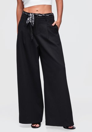 Femme portant un pantalon noir à jambes larges avec une ceinture foulard noir et blanc, un top court blanc et des talons ouverts noirs.